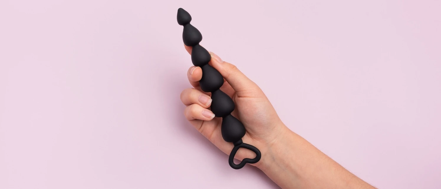 на рожевому фоні зображена рука, в якій силіконова чорна анальна пробка