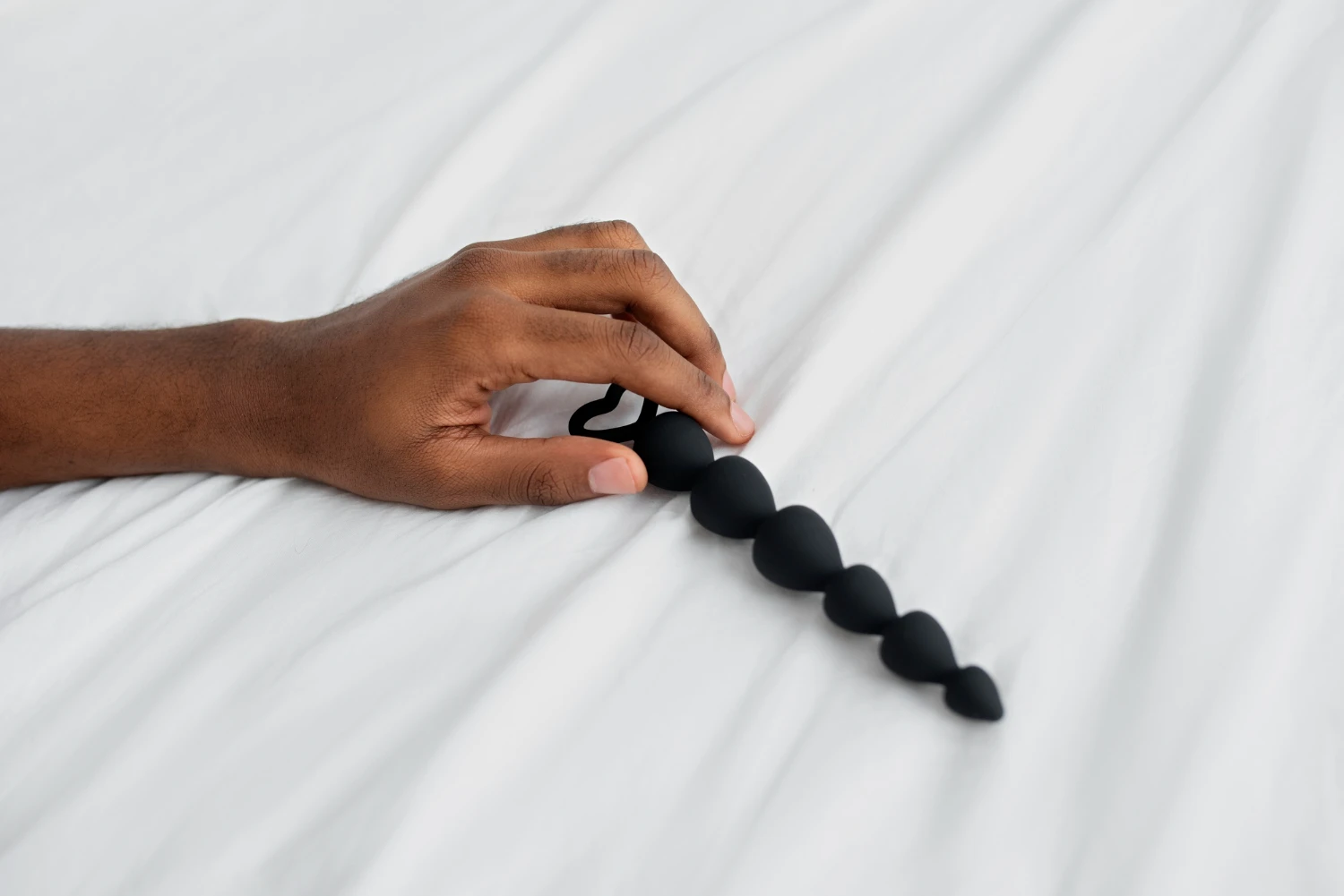 на белой простыни черная мужская анальная пробка из силикона в мужской руке