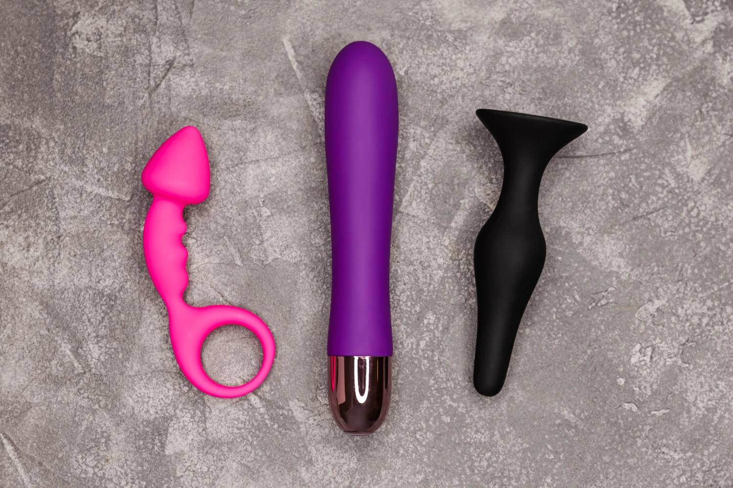 на сером столе лежит черная и розовая анальные пробки и анальный вибратор