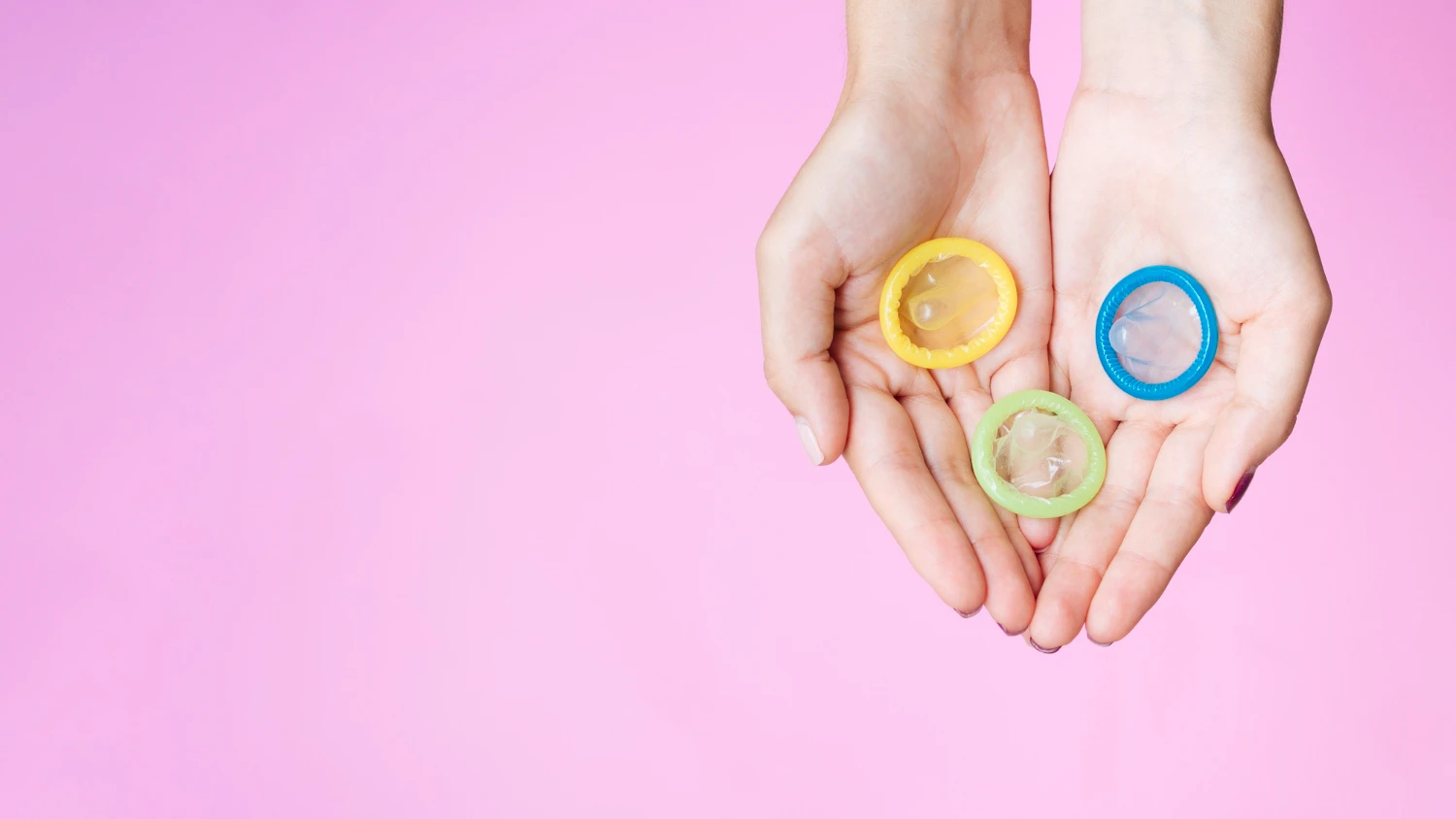 на картинке изображены распечатанные презервативы цветные в женской руке на розовом фоне