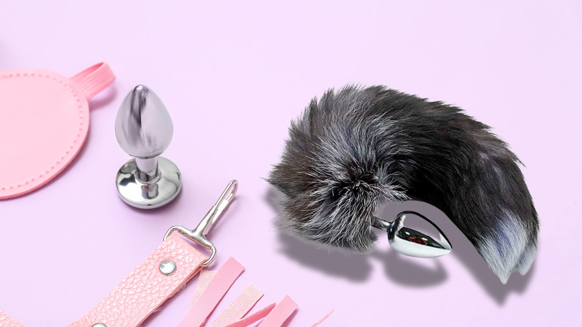на сиреньовому фоні зображена анальна пробка з хвостом, проста металева анальна втулка та рожеві аксессуари БДСМ