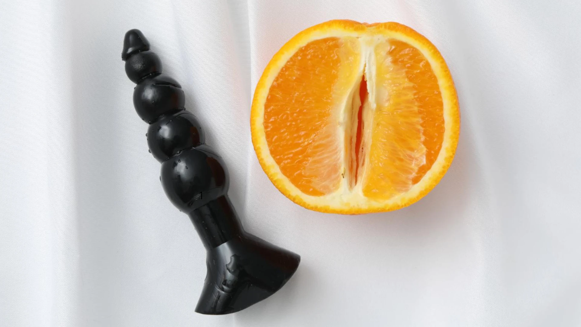 на фото велика черна анальна пробка та розрізаний апельсин на білій тканині