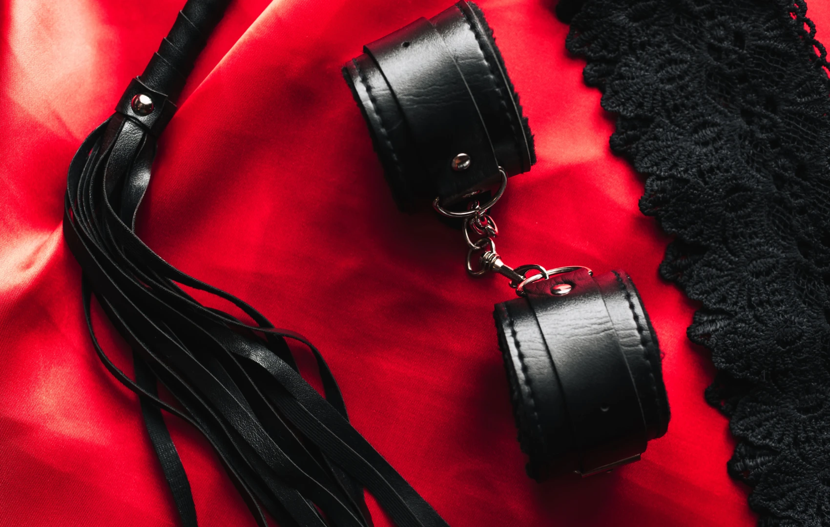 на красном шелке лежат черные наручники для БДСМ и черная плетка