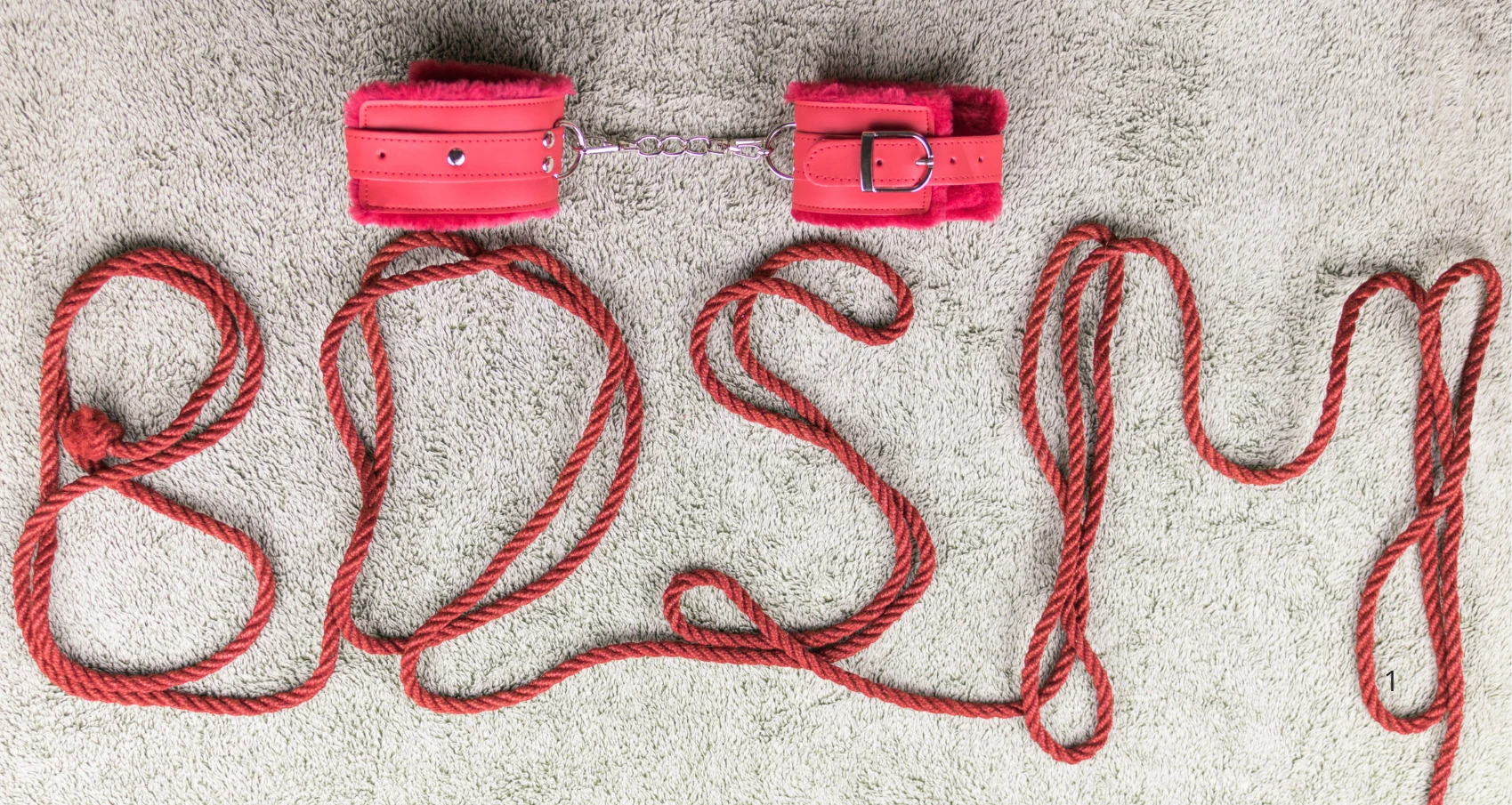на сером ковре лежат красные наручники для бдсм с мехом и веревка шибари
