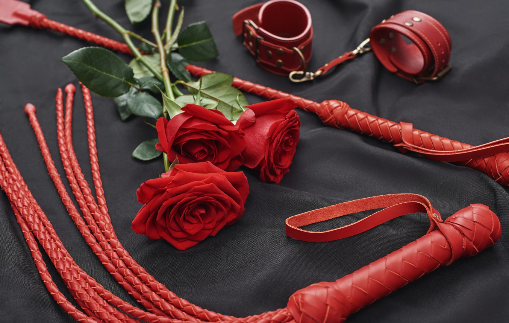 на чорному простирадлі червоний набір БДСМ: наручники, батіг, стек та червоні троянди