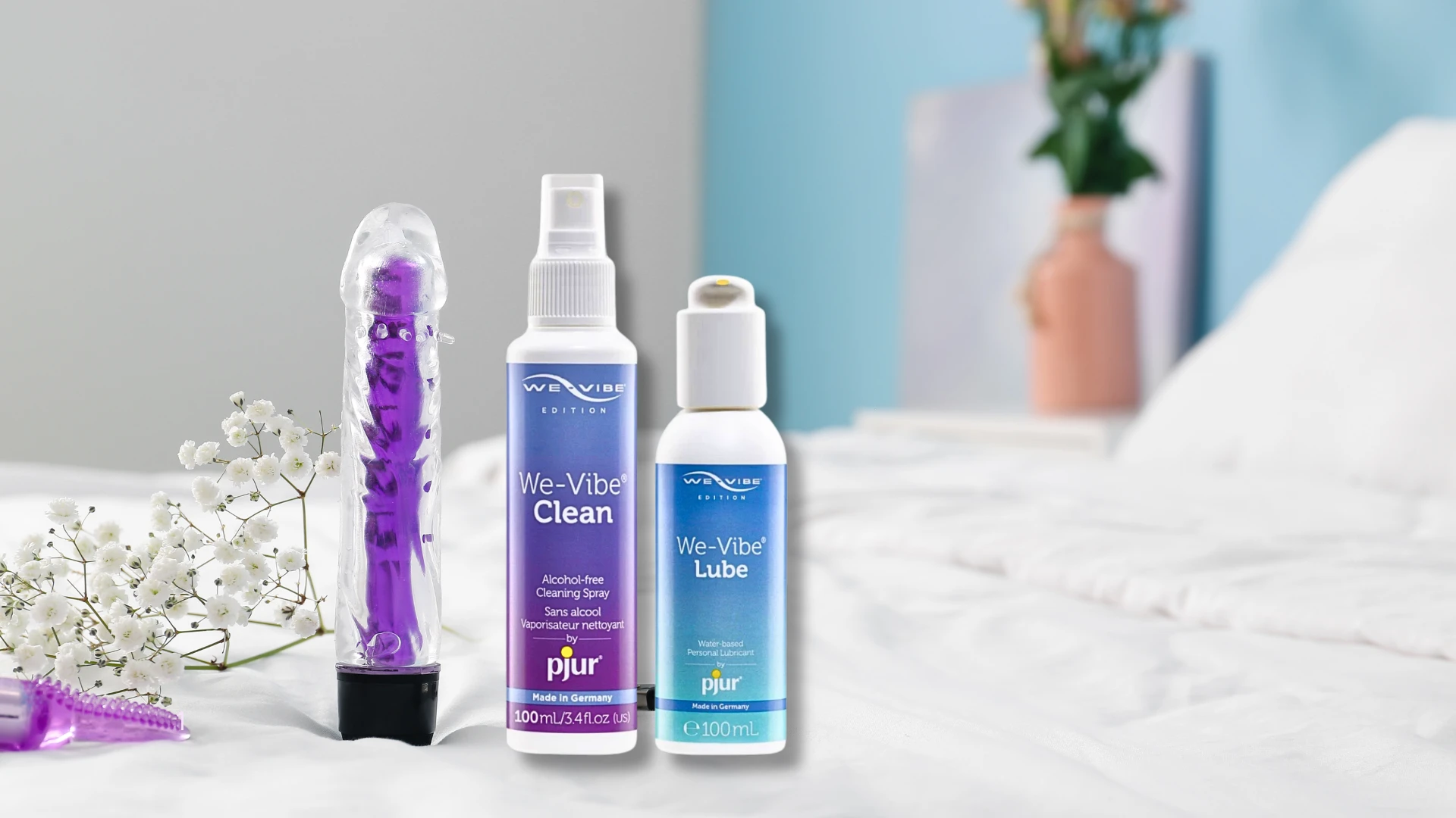на фото зображені очисники секс іграшок wi-vibe та вібратор на фоні ліжка з білим простирадлом