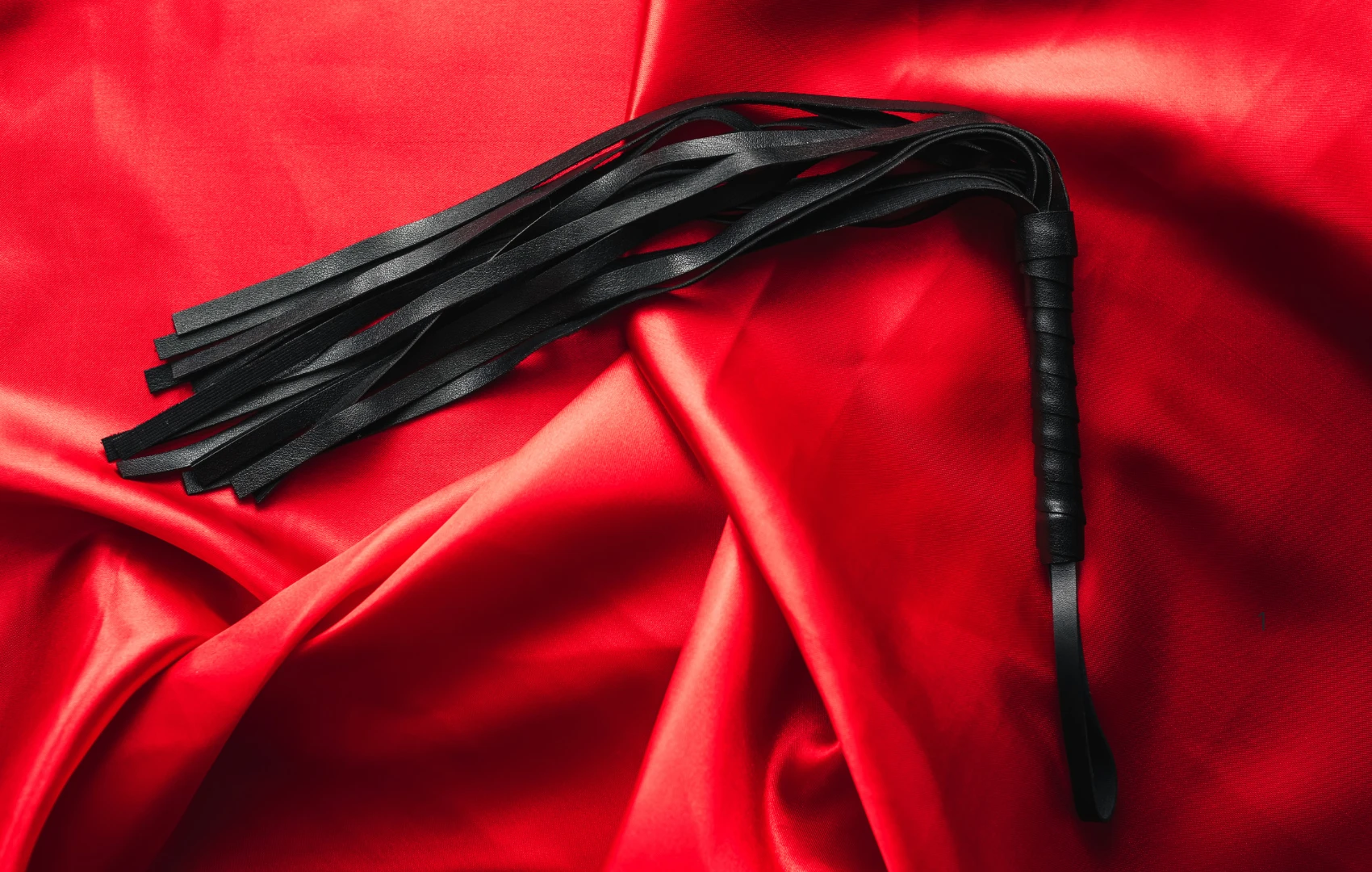 на червоному шовку лежить чорний батіг для БДСМ сексу
