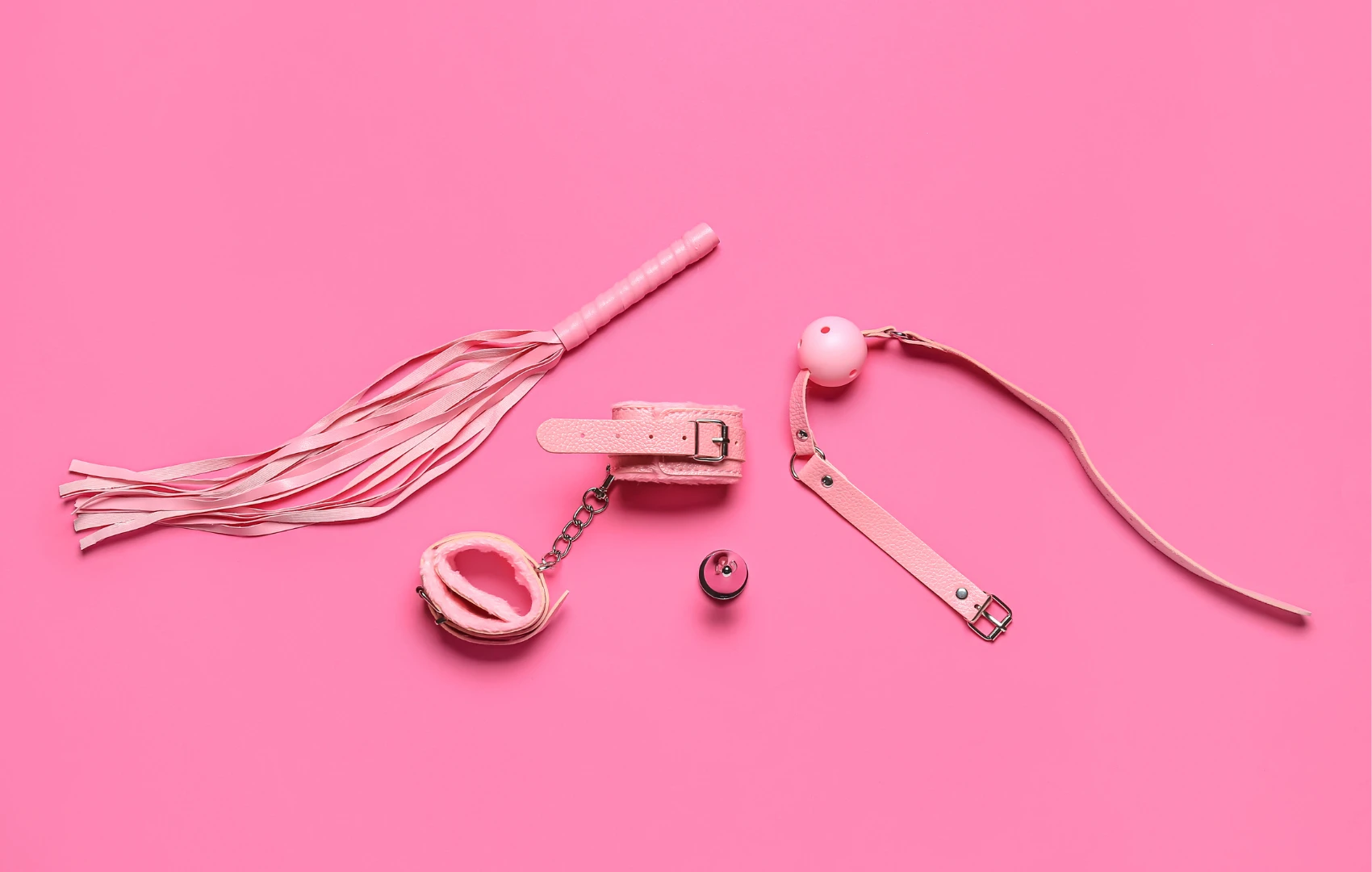 на рожевому фоні зображений рожевий батіг для сексу, рожевий кляп та рожеві наручники