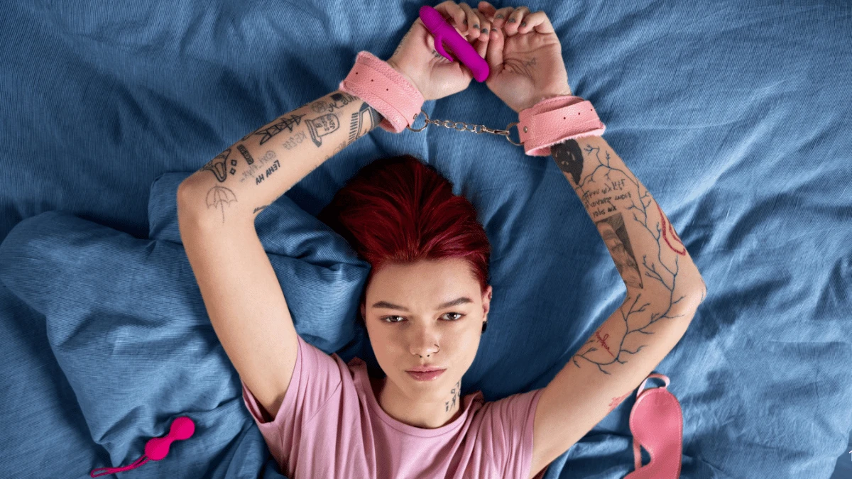 жіноча рожева бдсм маска, наручники рожеві та вагінальні кульки поряд з жінкою в ліжчі на блакитному простирадлі