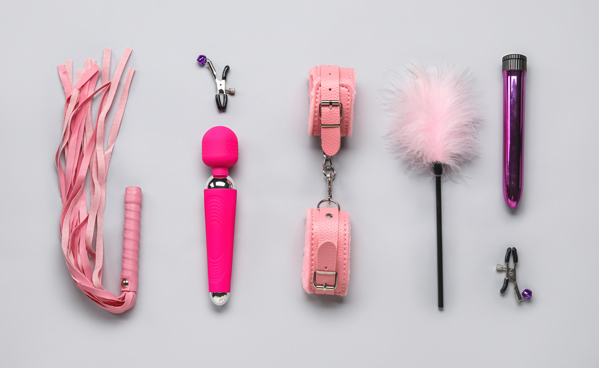 на сером фоне вибратор, розовые наручники и плетка, а также другие сексигрущки для женщин